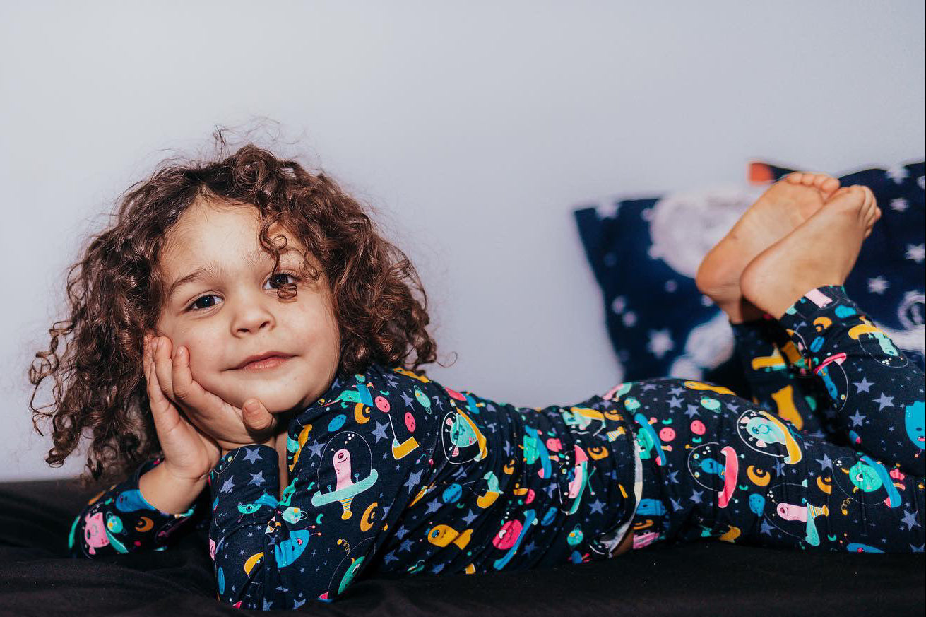 A young child with curly brown hair lies on a dark surface wearing a long-sleeve pajama top and matching pants with a colorful space-themed print of planets, stars, and aliens on a black background. 
